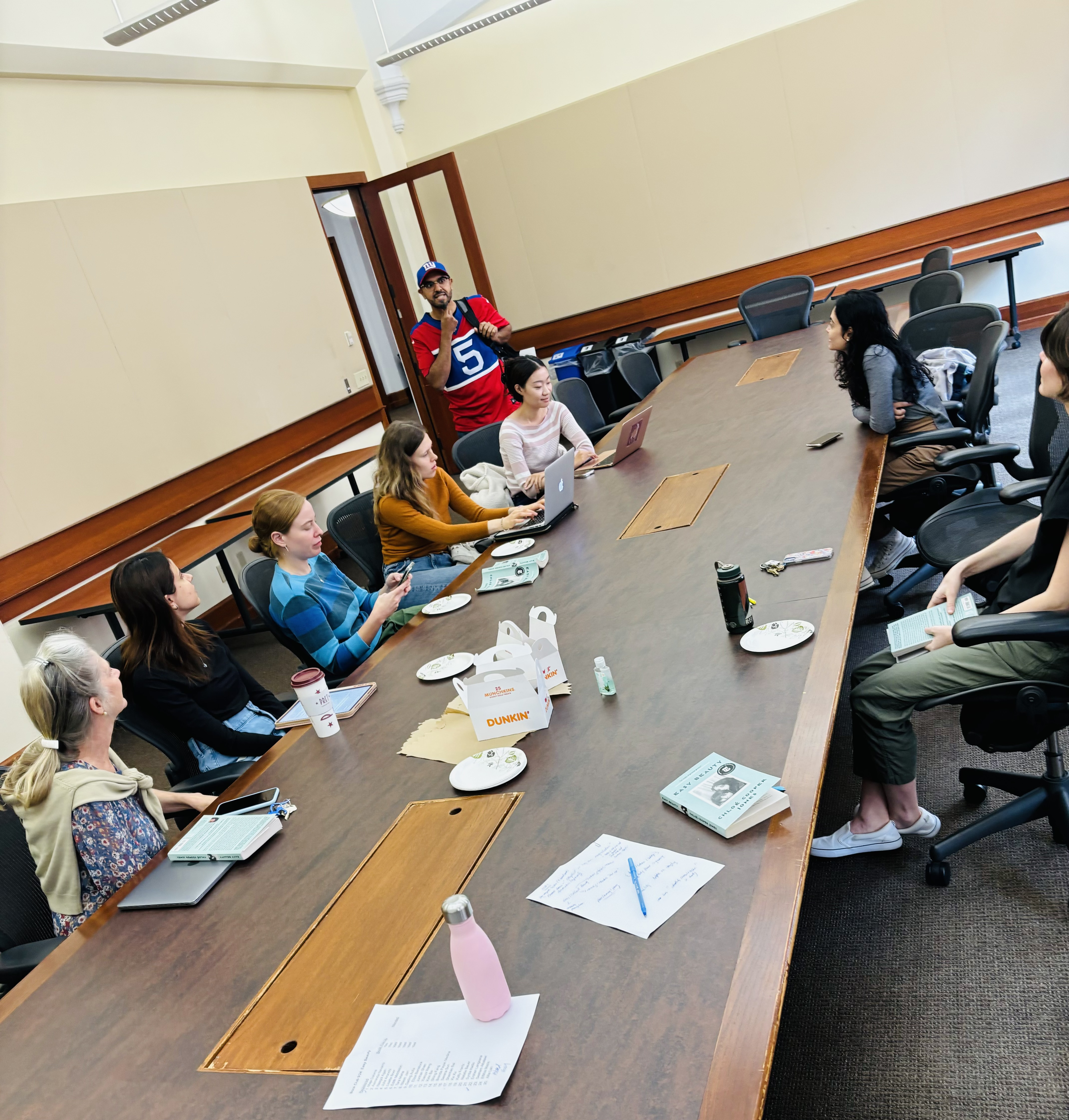 A group of people is gathered in a conference room around a large, rectangular wooden table. Most of the individuals are seated, using laptops, phones, and notebooks, while one person is standing in the background near the door wearing a red sports jersey and a blue cap. There are Dunkin' Donuts boxes, empty plates, and coffee cups on the table, suggesting a casual meeting or study session. The ro