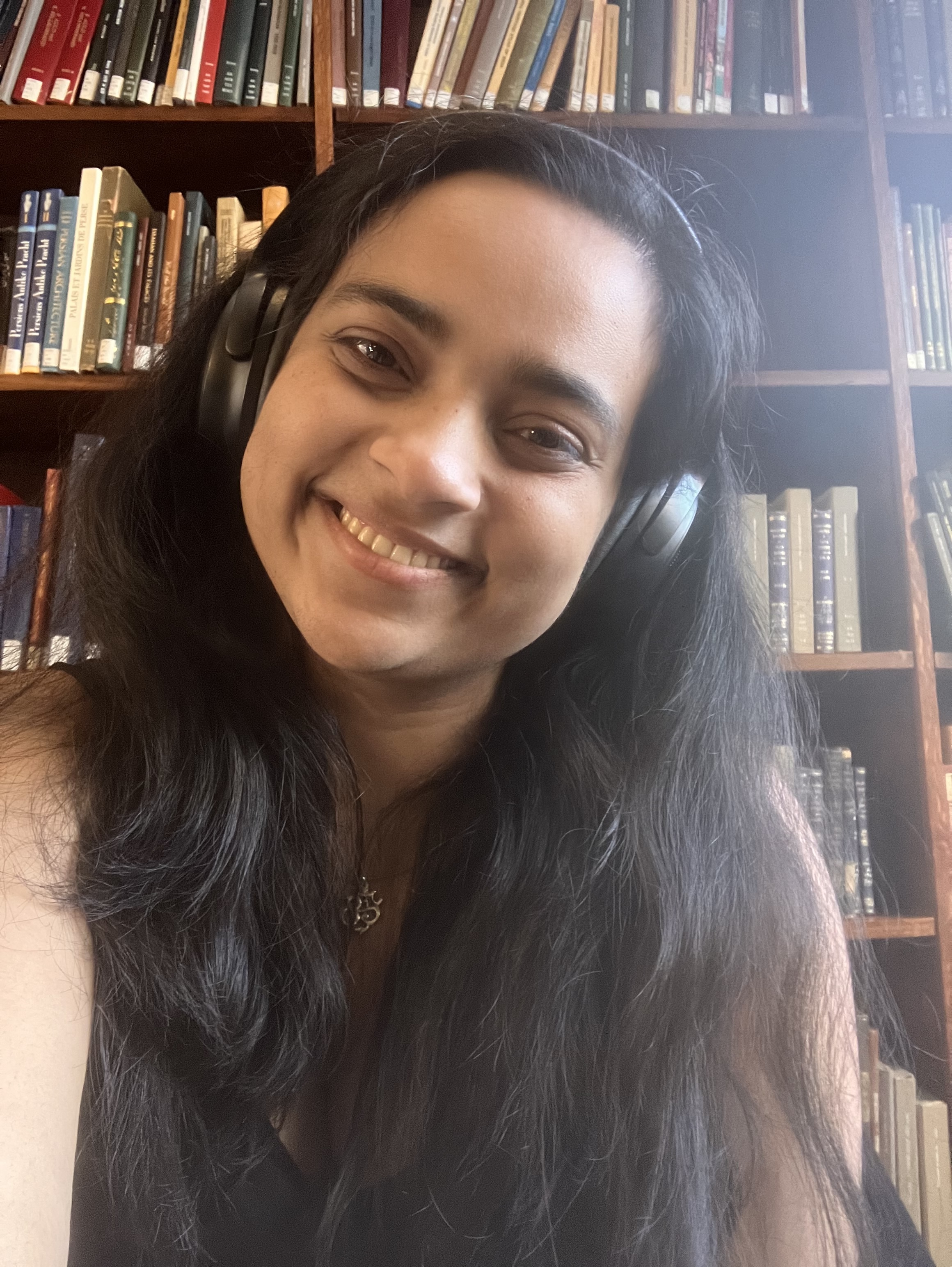A close-up selfie of a woman with long, dark wavy hair and dark brown eyes, wearing black over-ear headphones and a silver pendant necklace. She is tilting her head slightly with a broad smile. In the background, tall wooden library-style bookshelves are filled with rows of books.