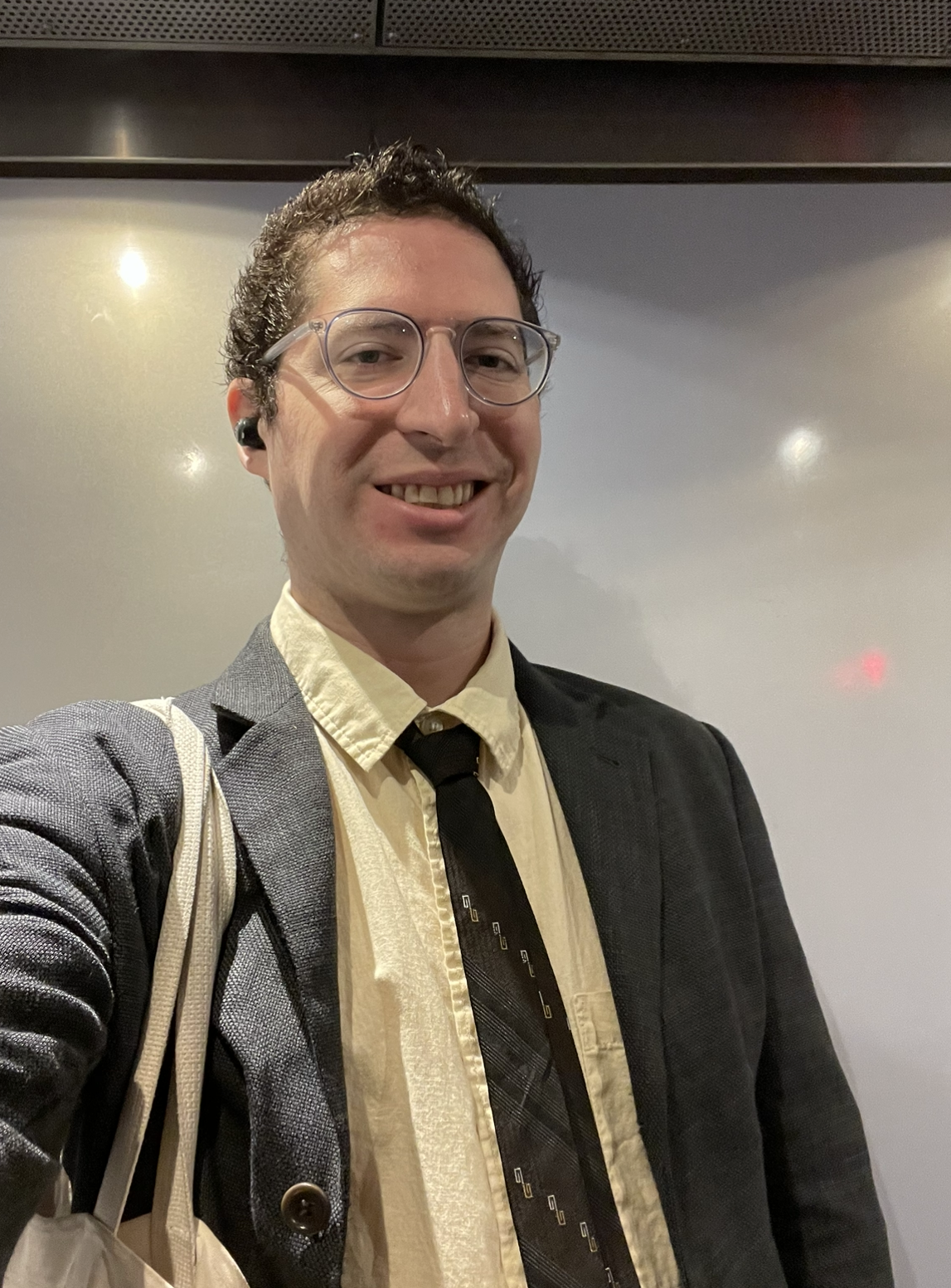 Headshot of a smiling man (Joel, he/him) dressed in a suit jacket and tie wearing glasses.