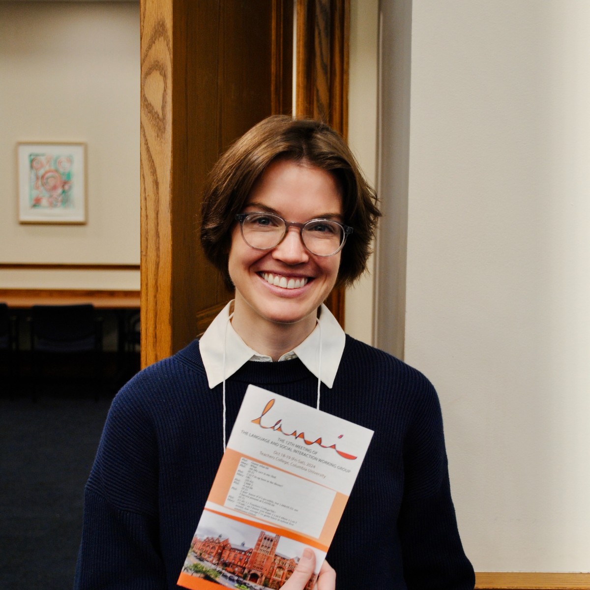 A smiling Kelly holding a pamphlet and posing in front of a classroom wall