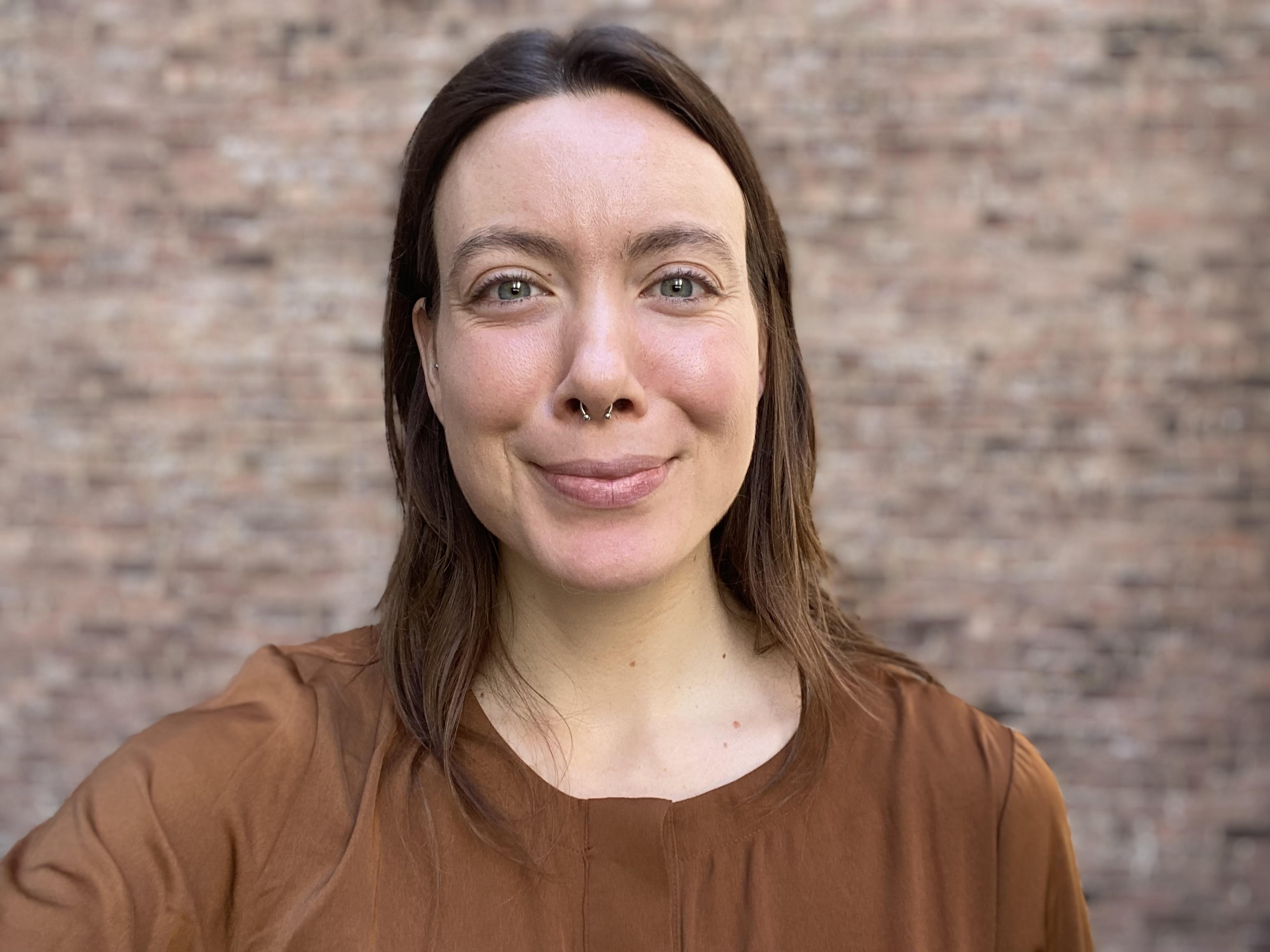 A photograph of a smiling Megan (she/her) wearing an earth tone colored shirt against a brick wall.