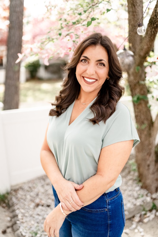 A woman with shoulder-length, wavy dark brown hair smiles warmly at the camera. She is standing outdoors in a garden with a flowering tree with pink blossoms behind her. She is wearing a sage-green v-neck blouse and dark blue jeans.