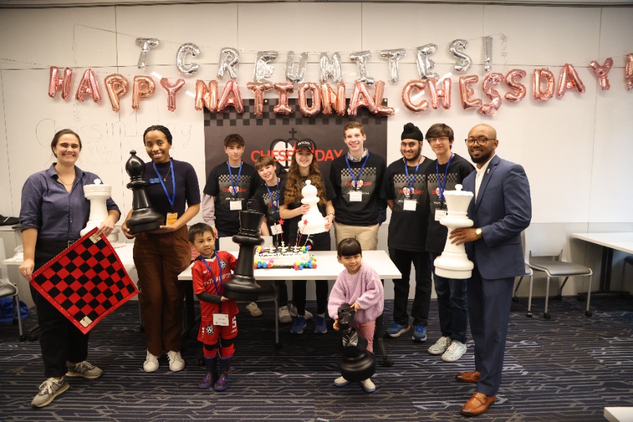 Group of adults and children posing in front of Chess Day signs holding chess boards and chess pieces