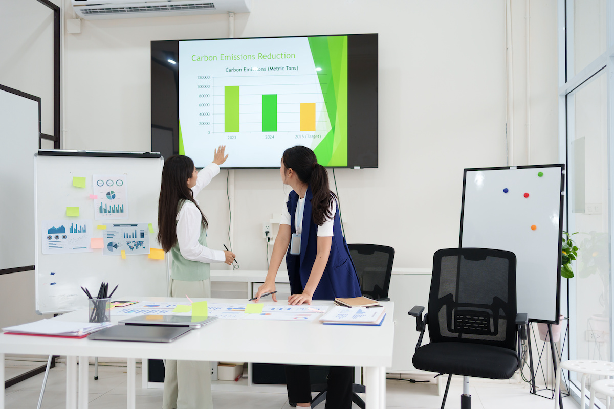 Two colleagues in a modern office setting looking at a TV screen behind them that shows a chart about carbon emissions reduction.