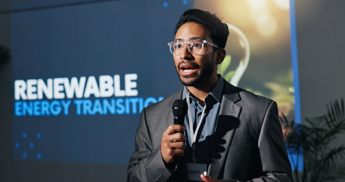 A male speaker with a microphone standing in front of a screen with the words renewable energy transition.