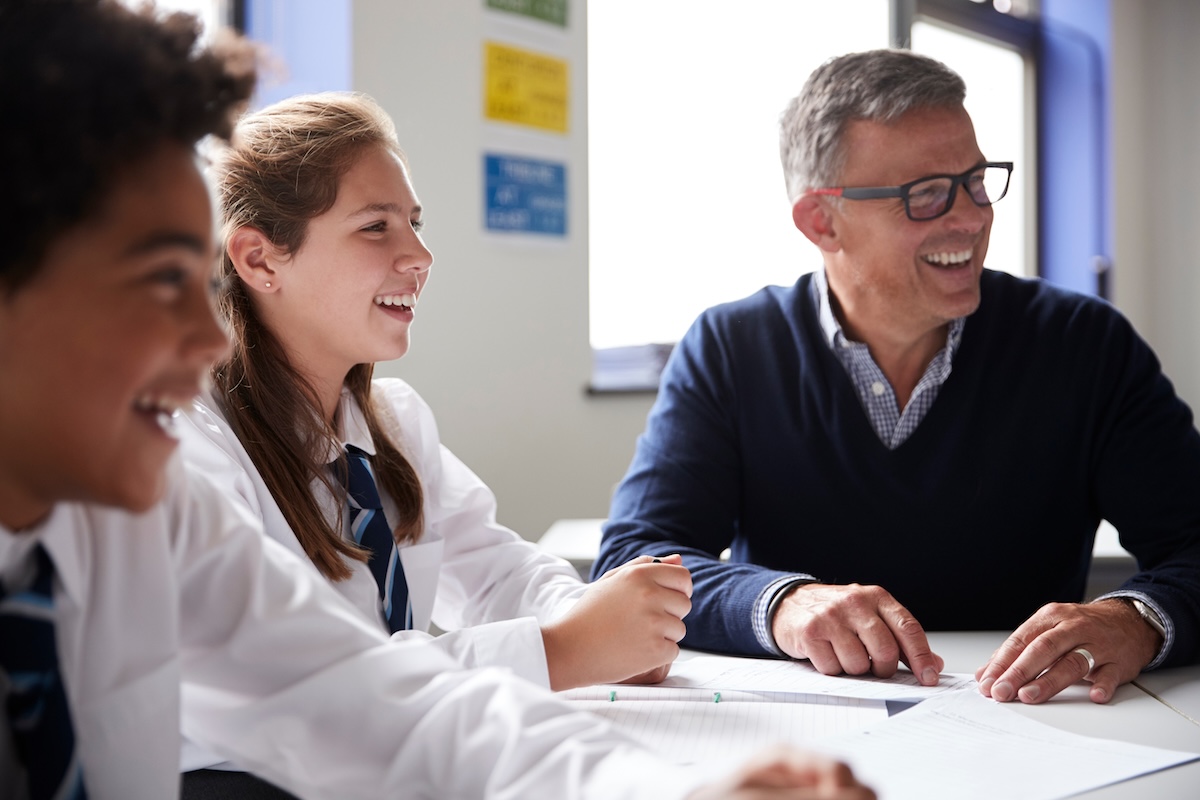 A man in professional attire sitting at a table with two children in school uniforms all smiling in a classroom setting