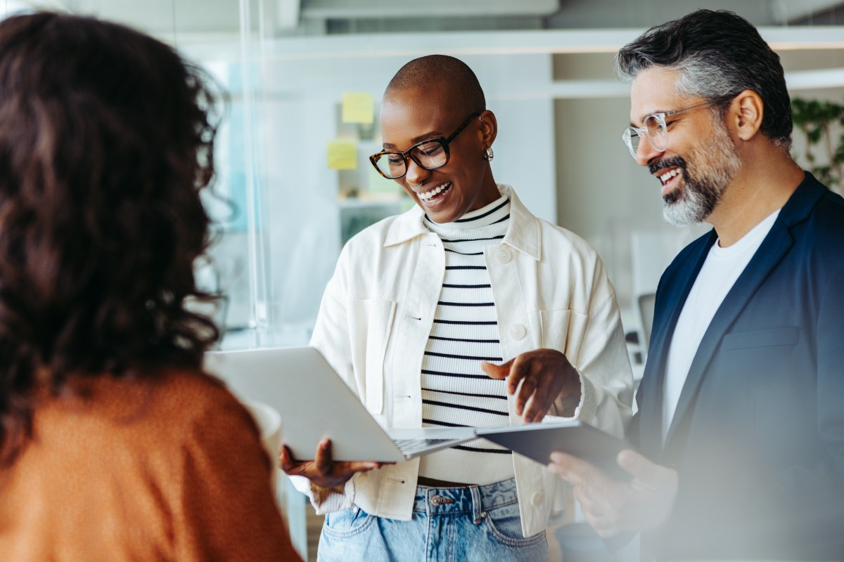A group of working professionals in an office setting standing together smiling while holding a laptop and tablet