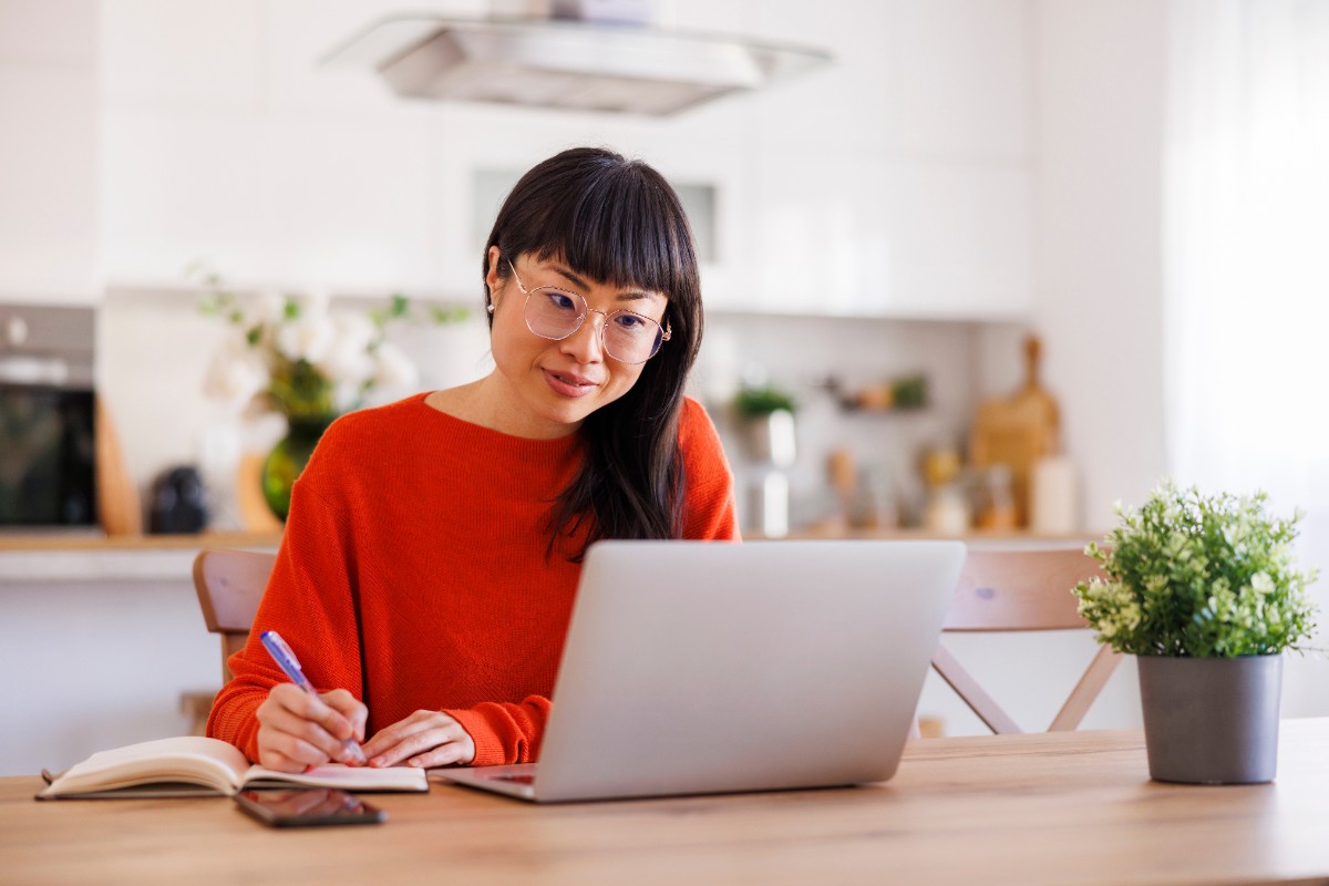 A woman sitting at a kitchen table writing in a notebook while looking at an open laptop