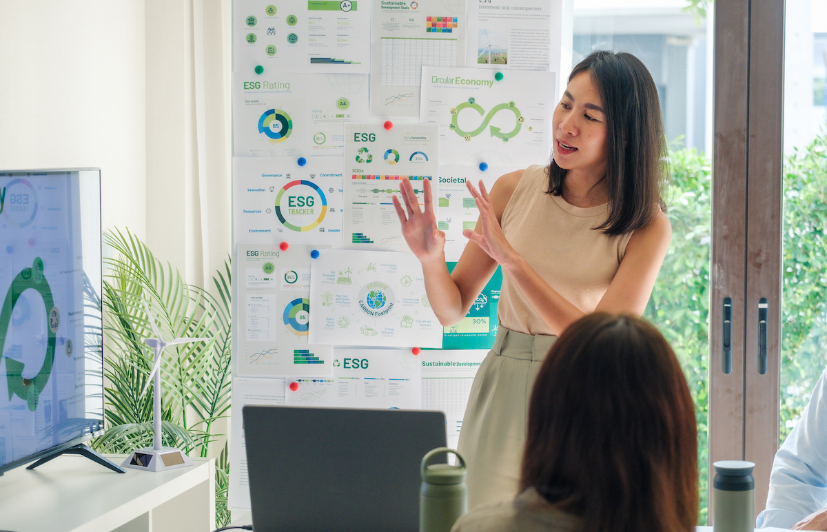 A woman giving a presentation in an office, standing in front of a wall covered with charts and graphs related to ESG (Environmental, Social, and Governance) topics, while two people listen.
