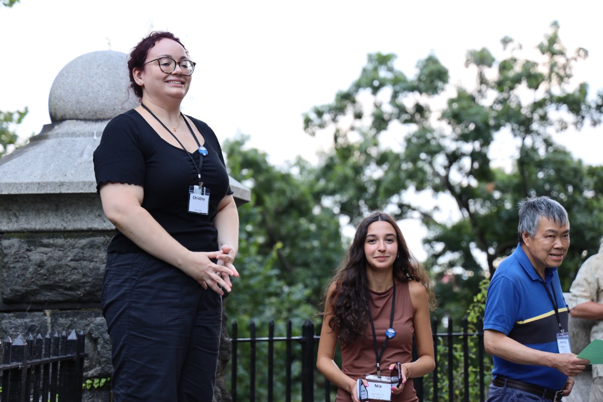 Three people outdoors wearing name tags, standing and sitting near a stone monument.