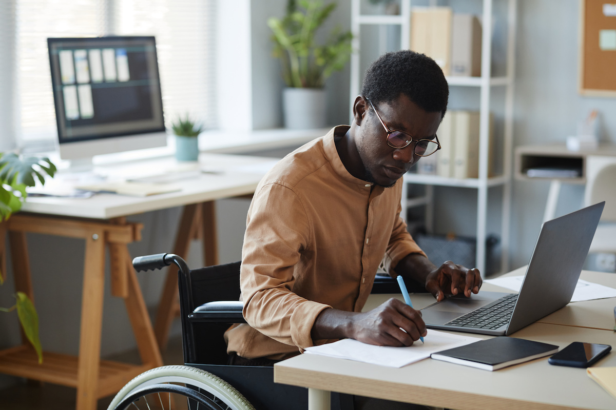 A man sitting at a desk working on a laptop and writing on a piece of paper with plants in the background