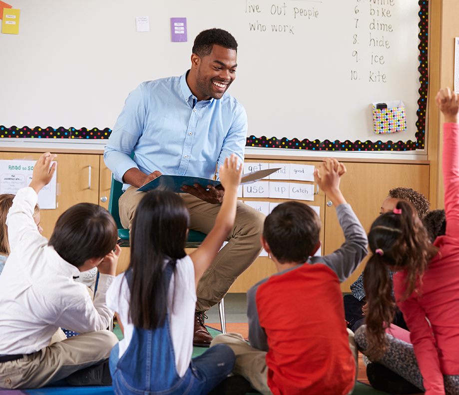 A teacher reading a book facing students with their hands raised