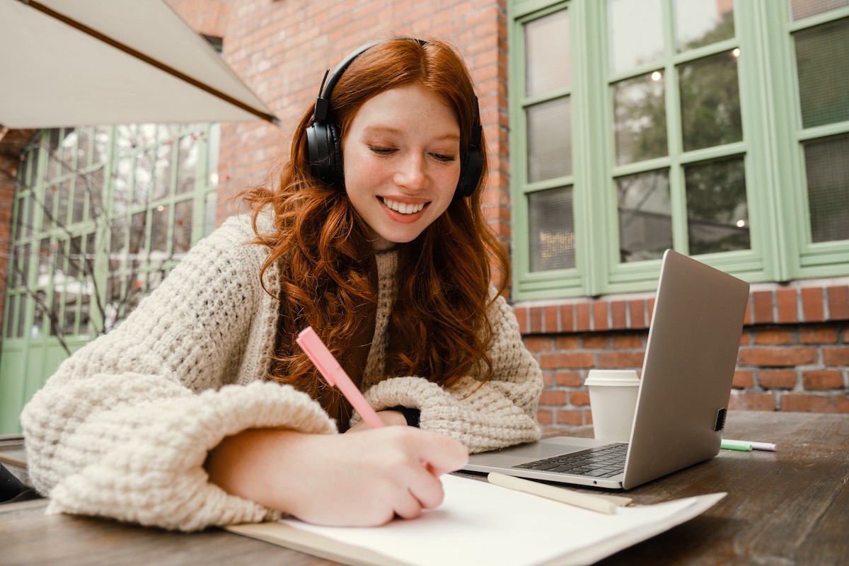 A woman wearing headphones sittiing on a bench outside writing in a notebook next to a laptop and a coffe cup