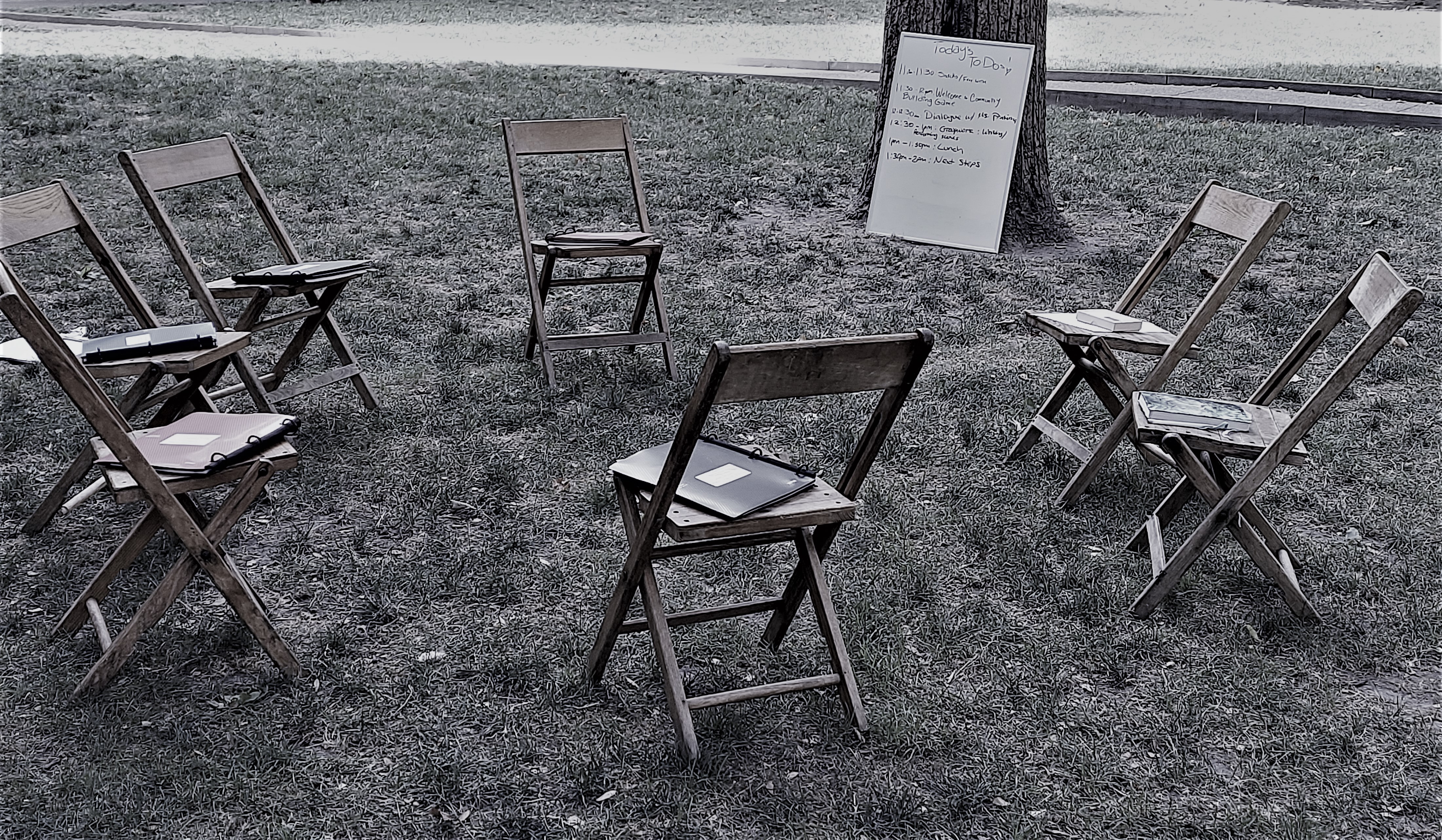 This is a black and white image of a circle of wooden folding chairs outside on the grass. On 5 of the chair seats are 3-ring binders, and on the 2 remaining chair seats, there are books without any lettering on them. A tree is nearby, and leaning against it is a 2-foot by 3-foot dry marker board that says 