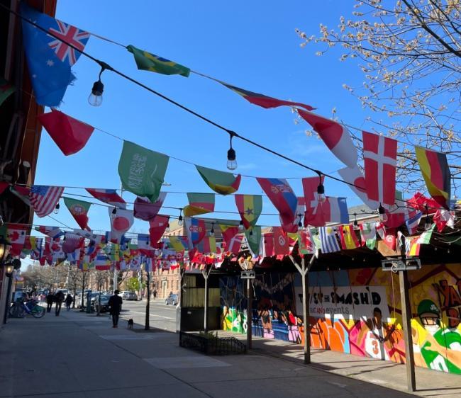 Streamers holding flags of countries from around the world wave above the sidewalk on a city street in Astoria, Queens