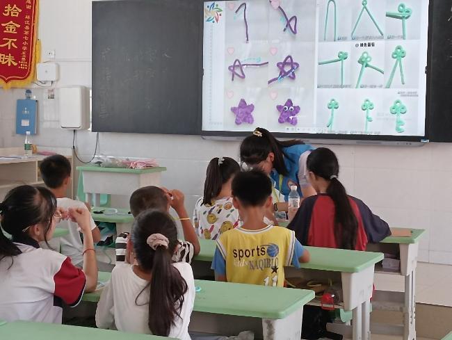 Children sit in rows in a classroom while an adult volunteer is at the front of the room teaching. A projector is showing the steps for knitting a star shape.