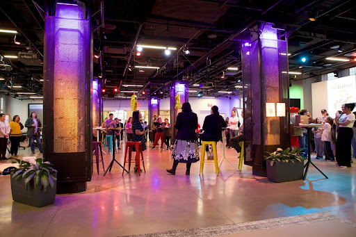 An image of MediaFest 2024 in the Smith Learning Theater. The Smith Learning Theater has an industrial look with colorful lighting and technology. There are many people mingling in the theater.