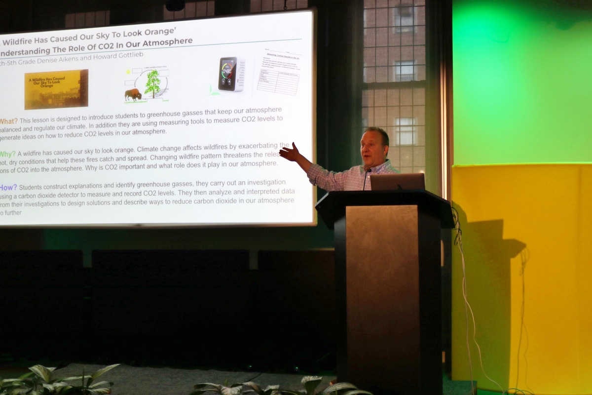 A white man gestures to a projector screen on the left side of the image while standing behind a podium. The screen is displaying text about a lesson plan developed around CO2 measurements