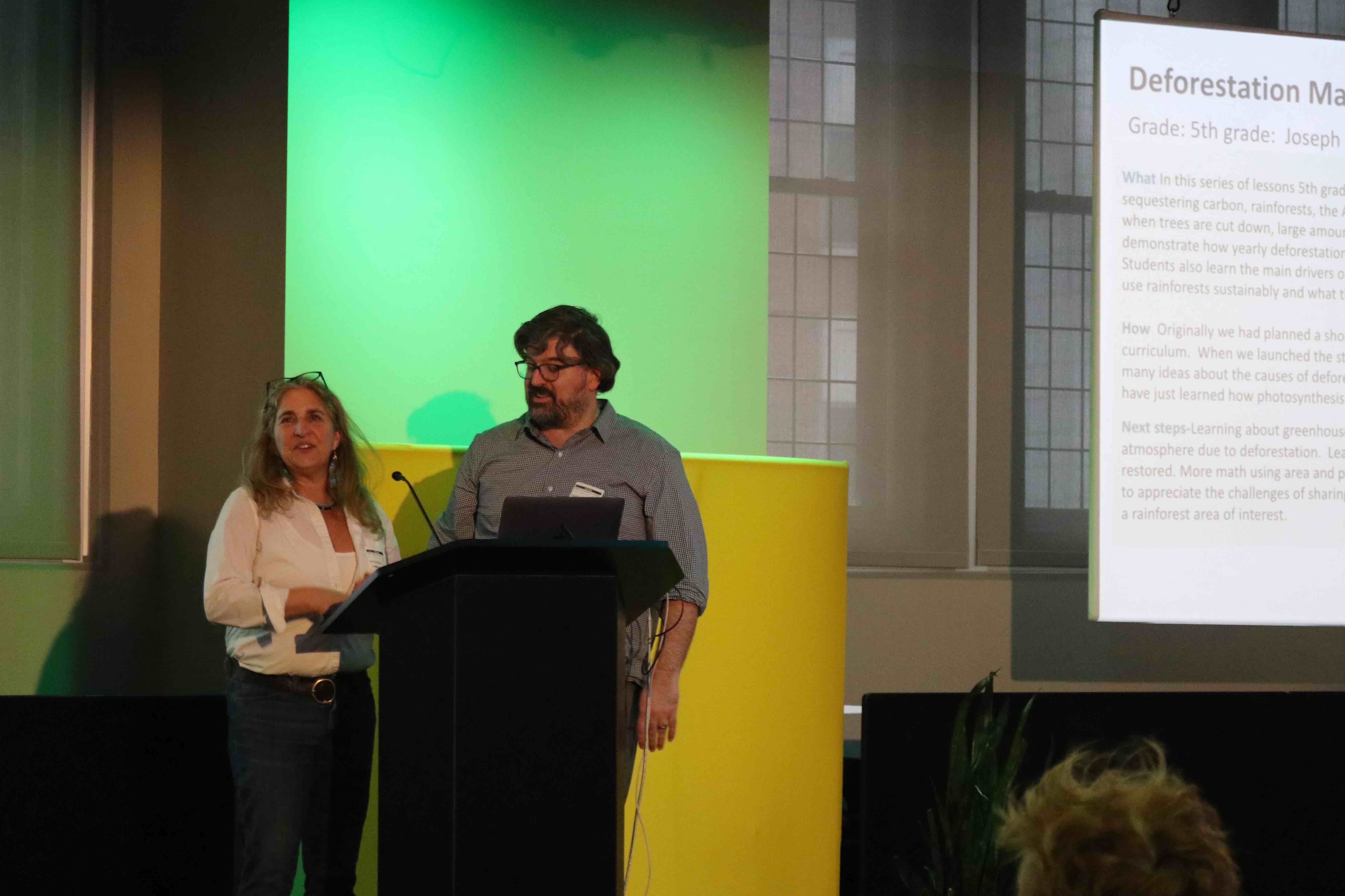 A woman and man stand next to each other giving a presentation. They are both standing behind a podium