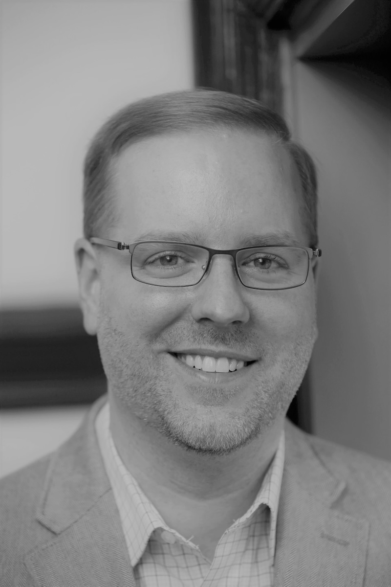 A black and white photo of Daniel Fienup smiling at the camera. He has fair skin, stubble and is wearing thin-rimmed glasses