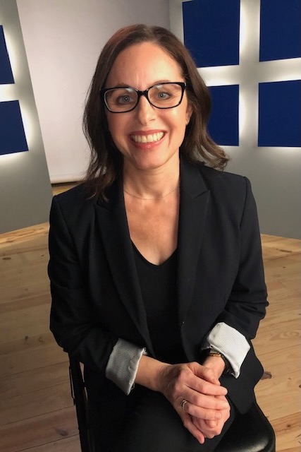 Randi Wolf smiling at the camera. She has shoulder length wavy brown hair, black glasses and is wearing a black blazer and black v-neck shirt.