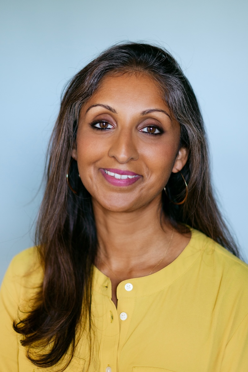 Sonali Rajan smiling at the camera. She has long gray-brown hair and brown skin and is wearing pink lipstick and a yellow blouse.