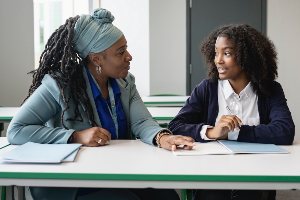 Front view of supportive Black female teacher in her early 50s sitting at desk with Black teenage schoolgirl while discussing her writing assignment.