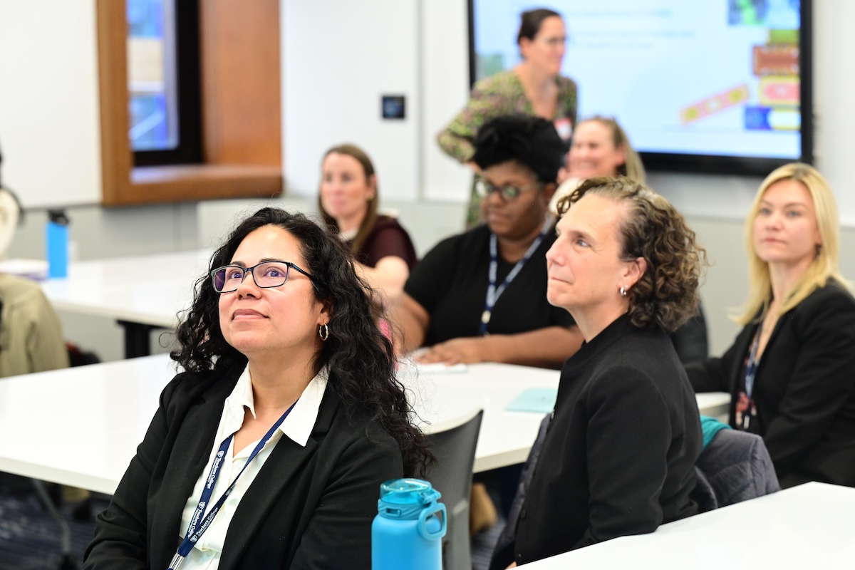 A group of women at a conference, sitting at tables and looking toward a speaker