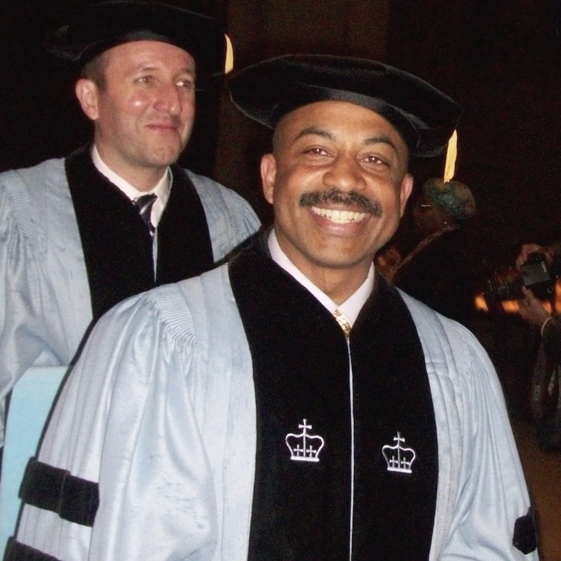 Dennis chambers at doctoral convocation. He his smiling and wearing Columbia blue doctoral robes