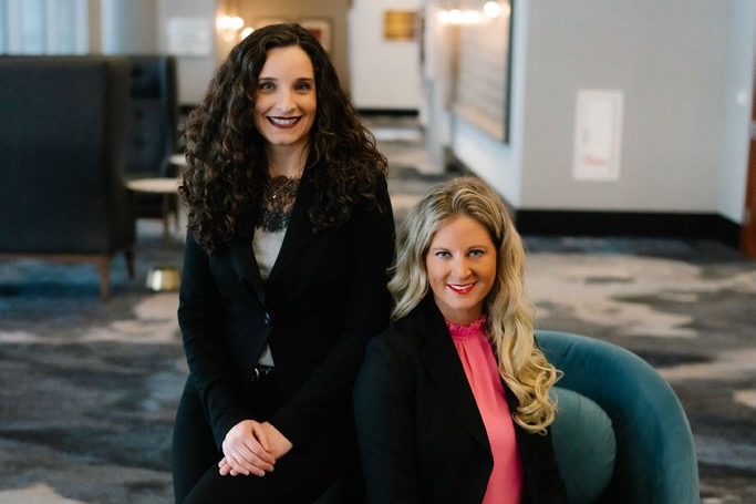 Diana Yassanye, with long dark hair and a dark suit, sits on the back of a chair next to Ranelle Brew, a woman with long blonde hair and black blazer and bright pink top