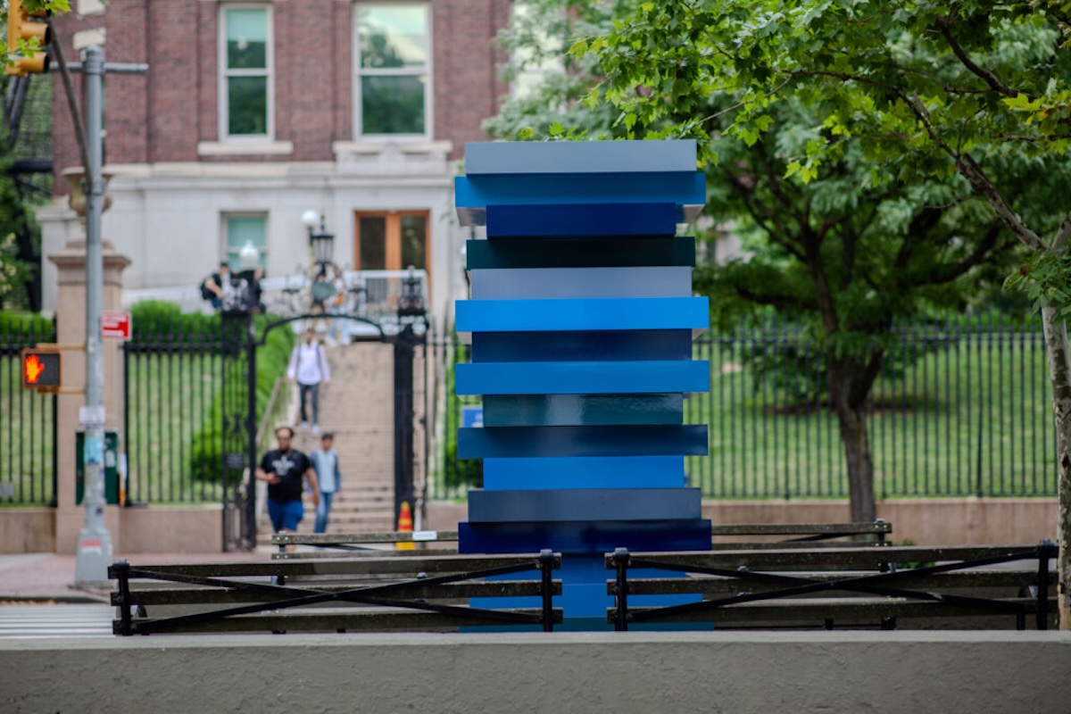 Sculpture outside a college building. It consists of at least 16 blue sheets of steel, in various shades, stacked atop each other