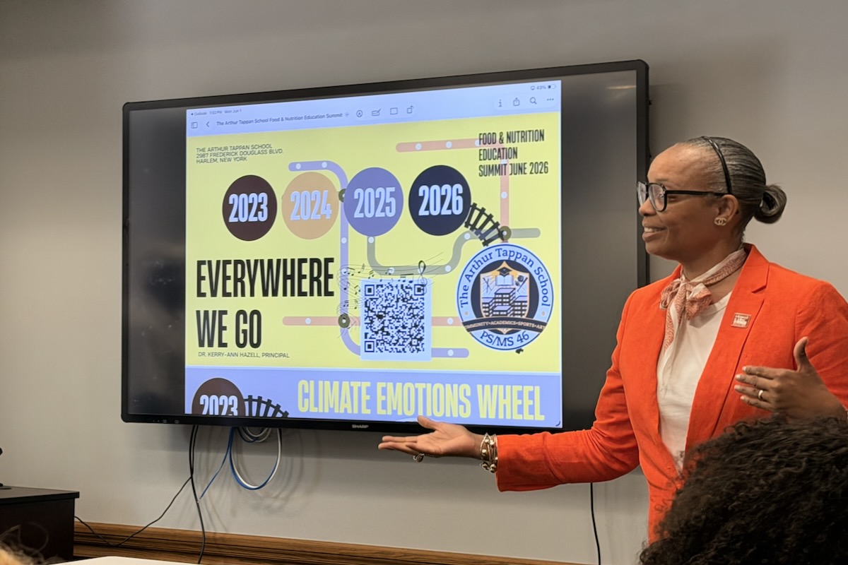 A Black woman in an orange blazer smiles infront of a projector with a slideshow presenation