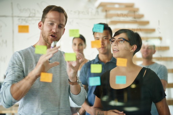 A group of colleagues standing behind a transparent whiteboard with writing and various colored Post-It notes on it