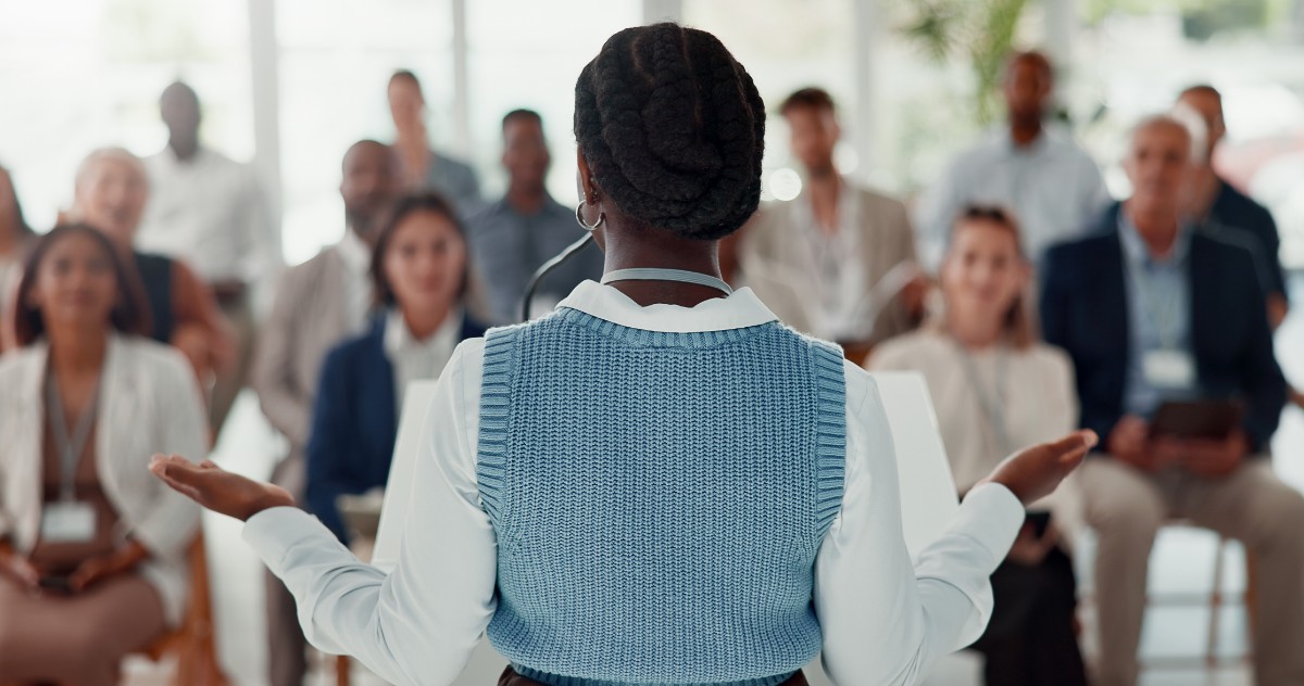 View of the back of a woman standing and speaking in front of a large group